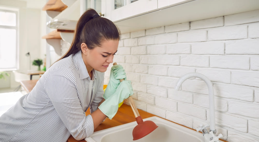 Woman having a problem with the sink. Young woman doing work about the house and trying to unblock the food remains clogged in the kitchen sink drain with a plunger.
